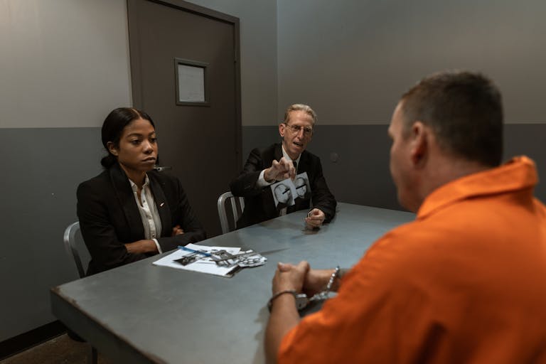 A suspect in an orange jumpsuit is interrogated by detectives in a dimly lit room.